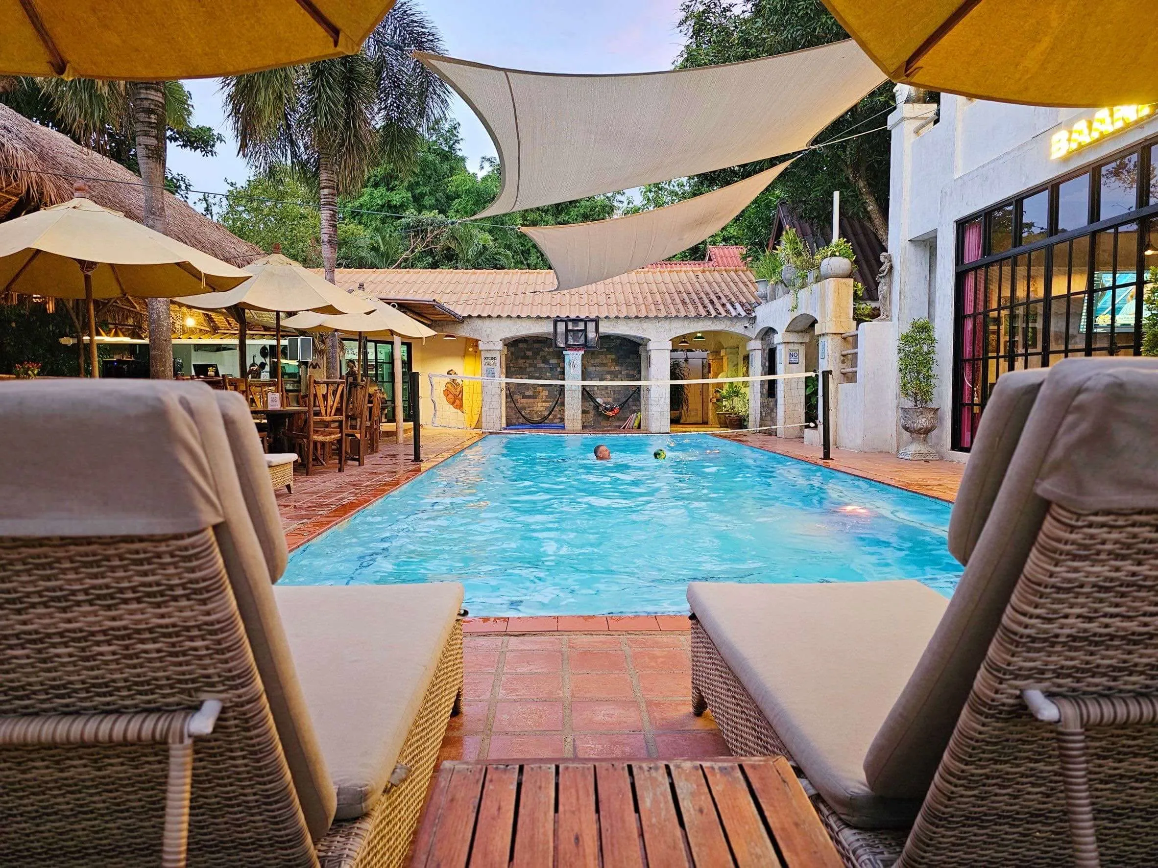 The pool at dusk seen from between two loungers, under the shade sails, with the restaurant lit behind it